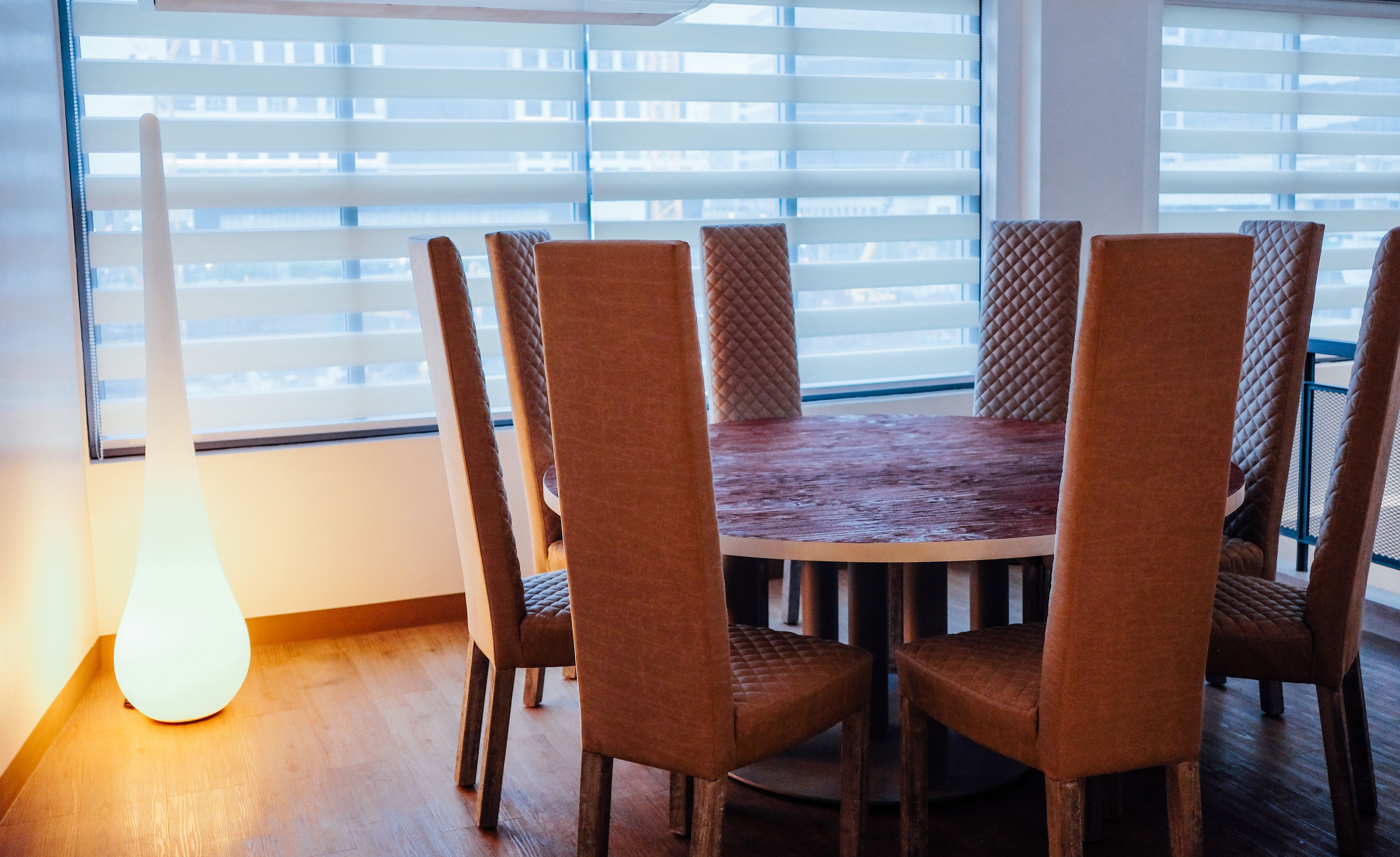 A dining area with a round table surrounded by six chairs, wooden flooring, and a window with blinds.