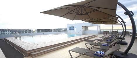 Un área de piscina en la azotea con sillones de descanso y sombrillas, una piscina y un pequeño edificio al fondo.