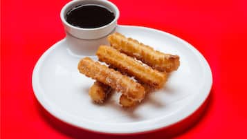 A plate of fried churros with a dipping sauce on a red background.