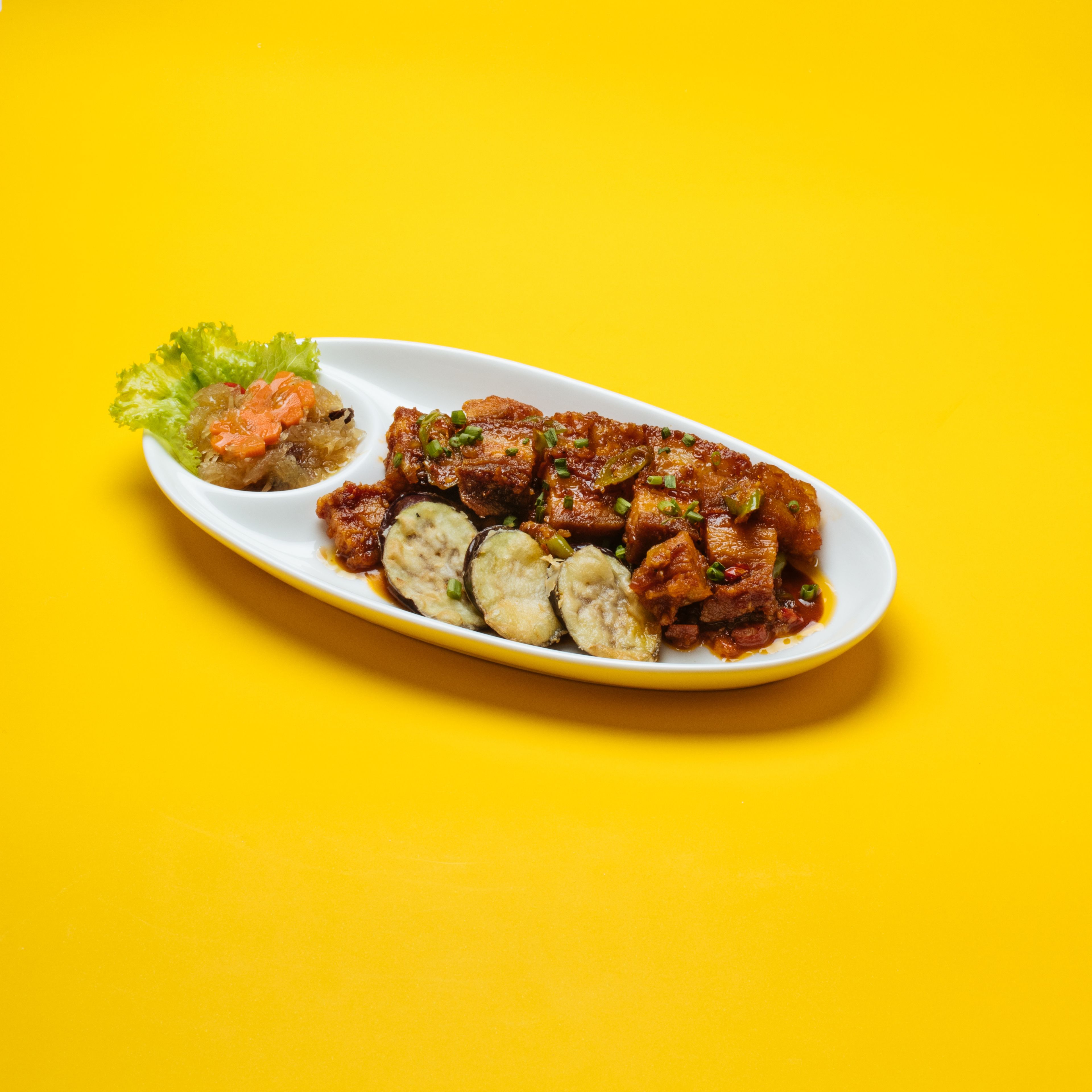 Un plato ovalado blanco con diversos alimentos, incluyendo carne en rodajas, verduras y una guarnición de vegetales encurtidos, sobre un fondo amarillo.