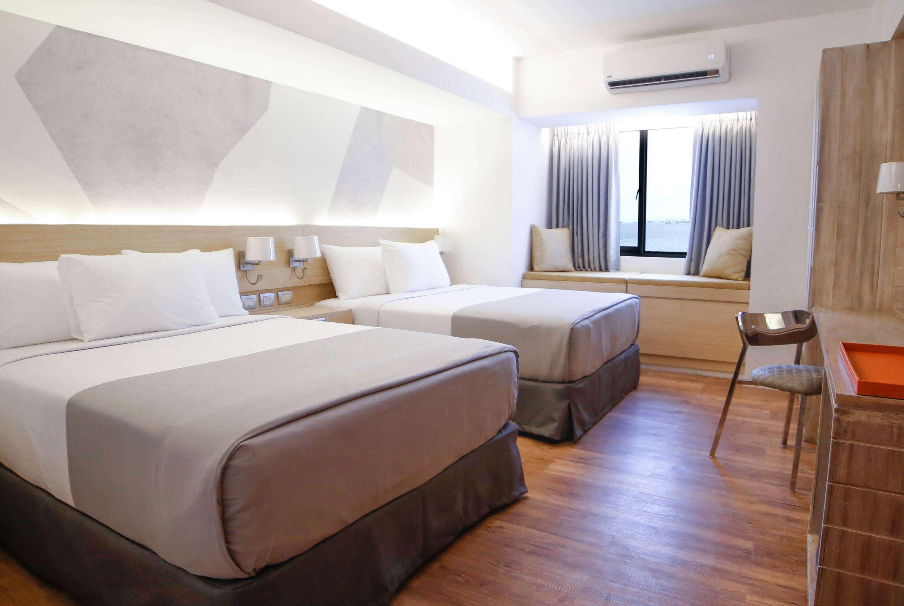 A hotel room with two beds, a window with curtains, an air conditioning unit, and a wooden desk with a chair.