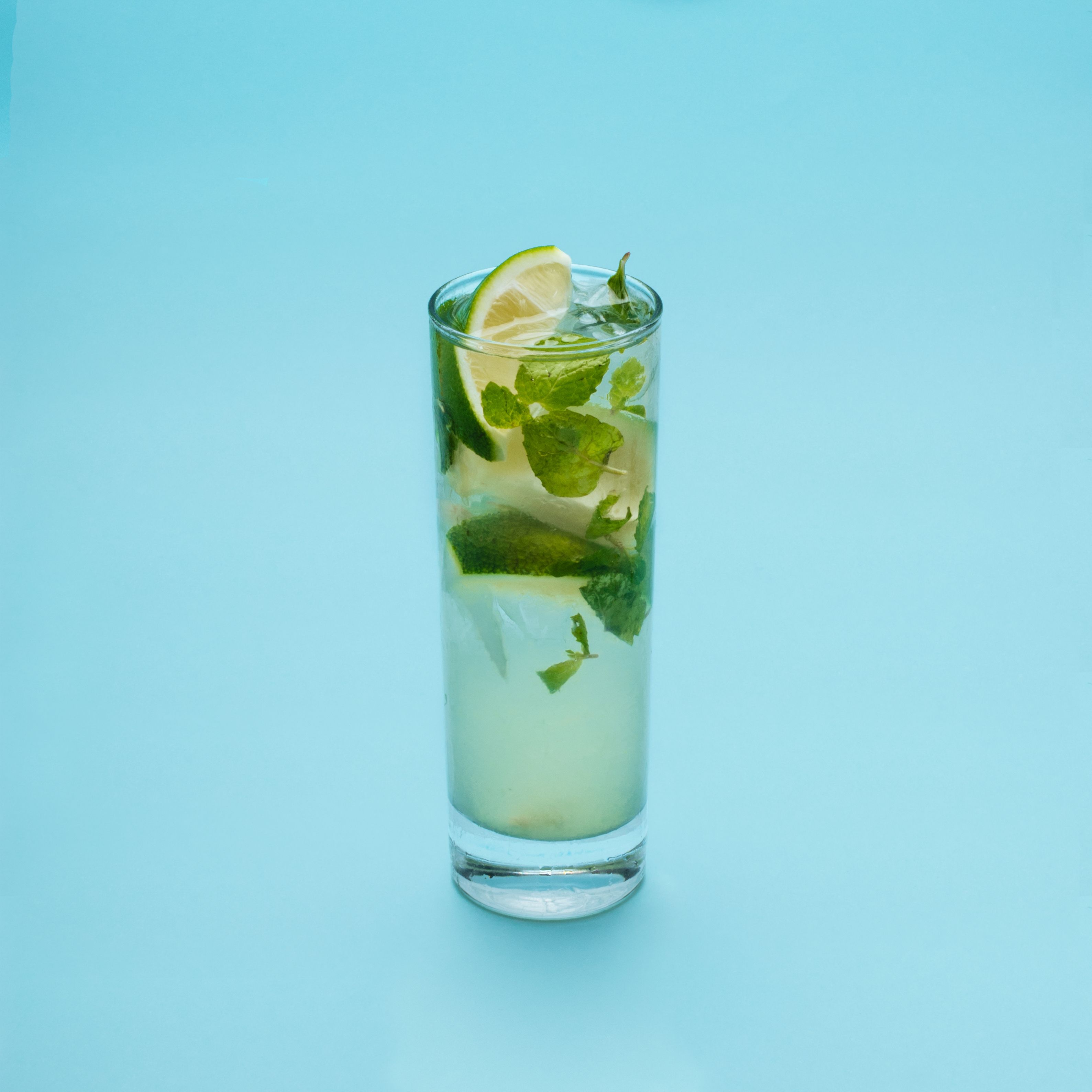 A tall glass with a lime slice and mint leaves filled with a clear liquid, set against a light blue background.