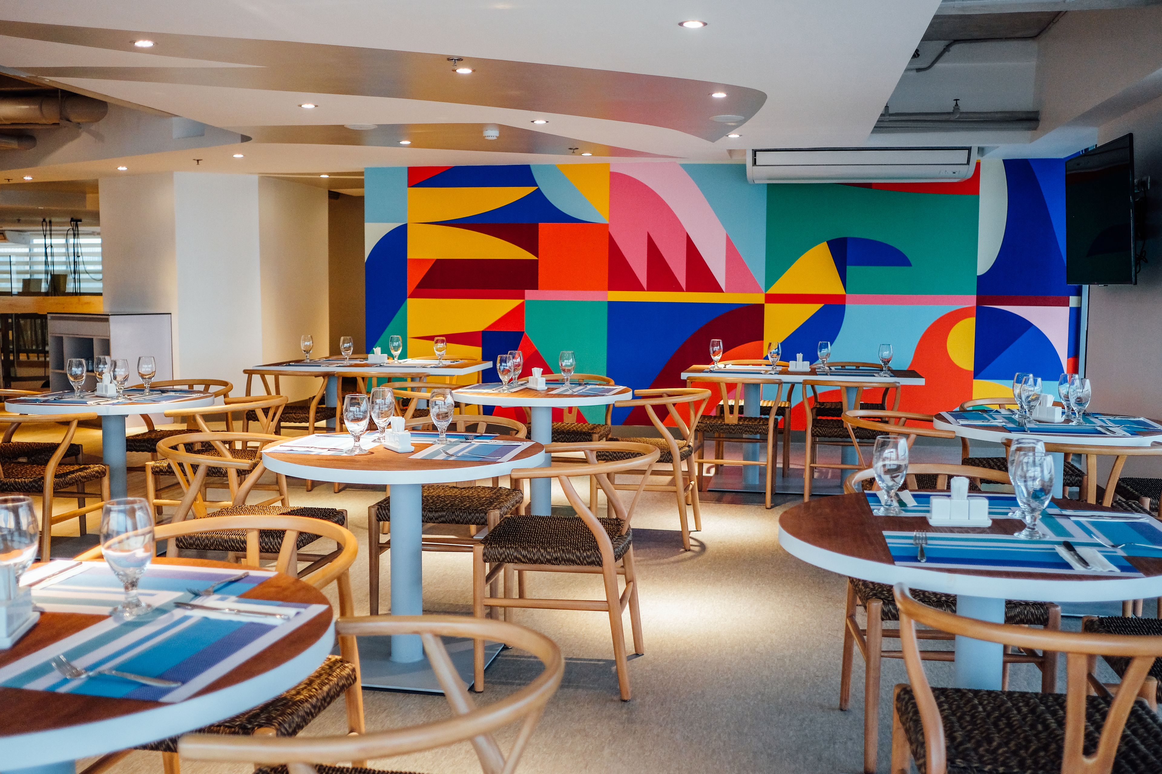 Un restaurant avec des tables rondes et des chaises en osier, agrémenté d’une fresque murale abstraite aux couleurs vives.