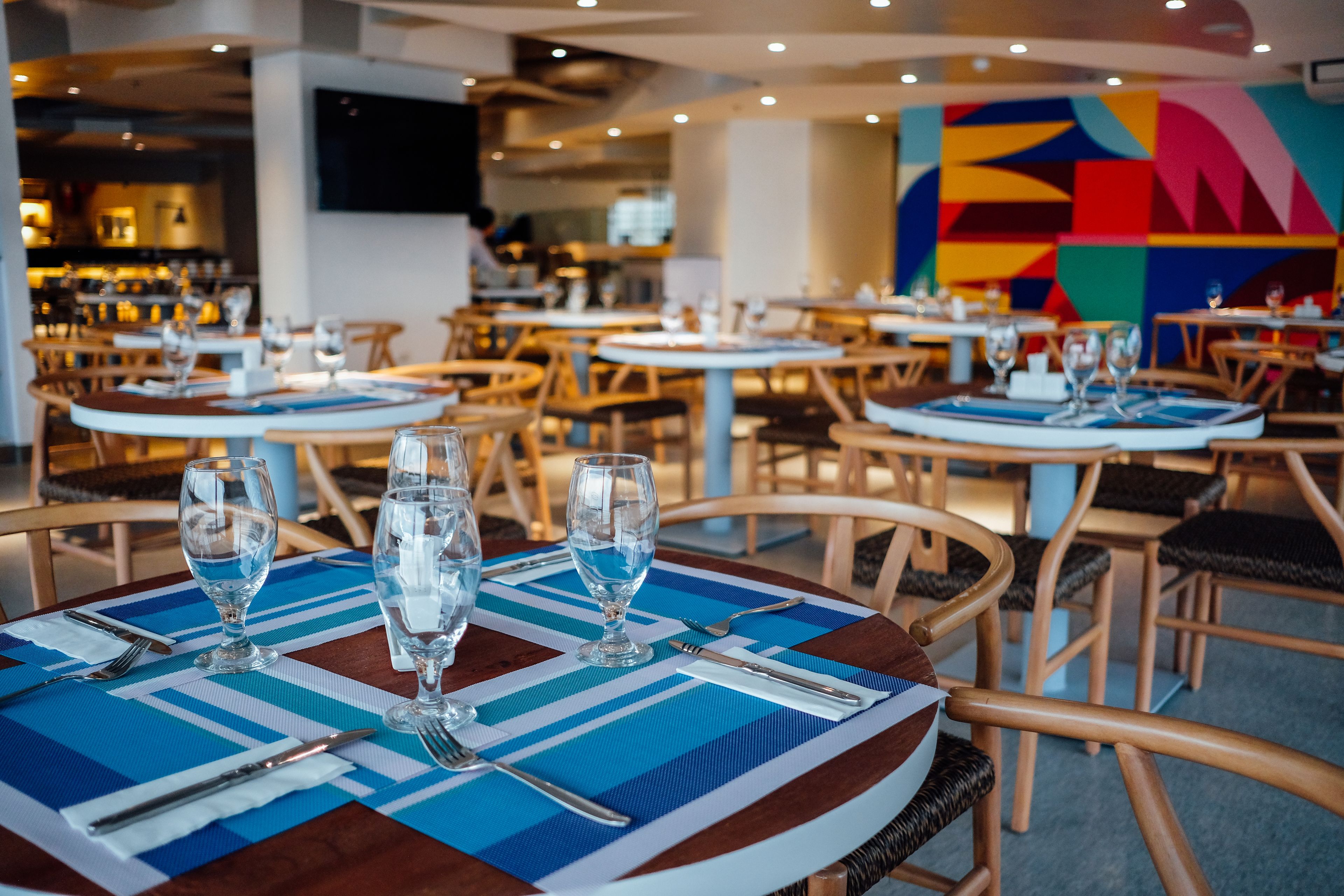 A restaurant with round tables set with blue placemats, silverware, and empty wine glasses.