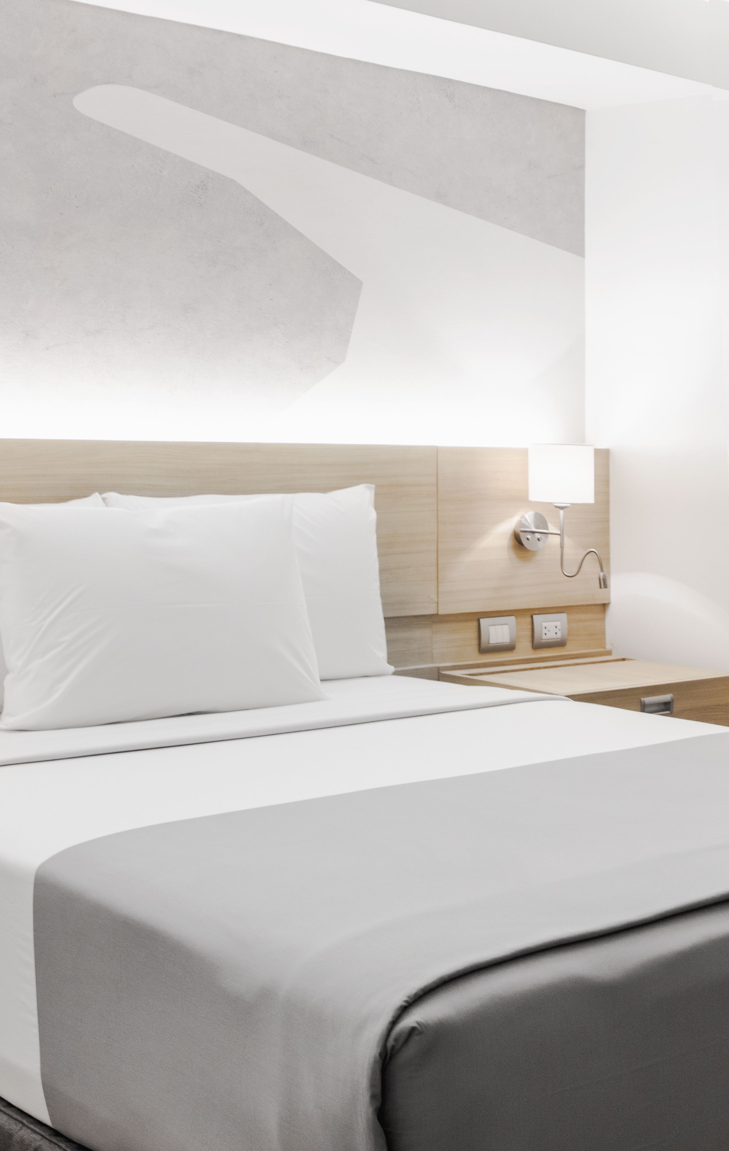 A modern hotel room with a neatly made bed, a bedside table with a lamp, and a wall-mounted light fixture.