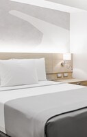 A modern hotel room with a neatly made bed, a bedside table with a lamp, and a wall-mounted light fixture.