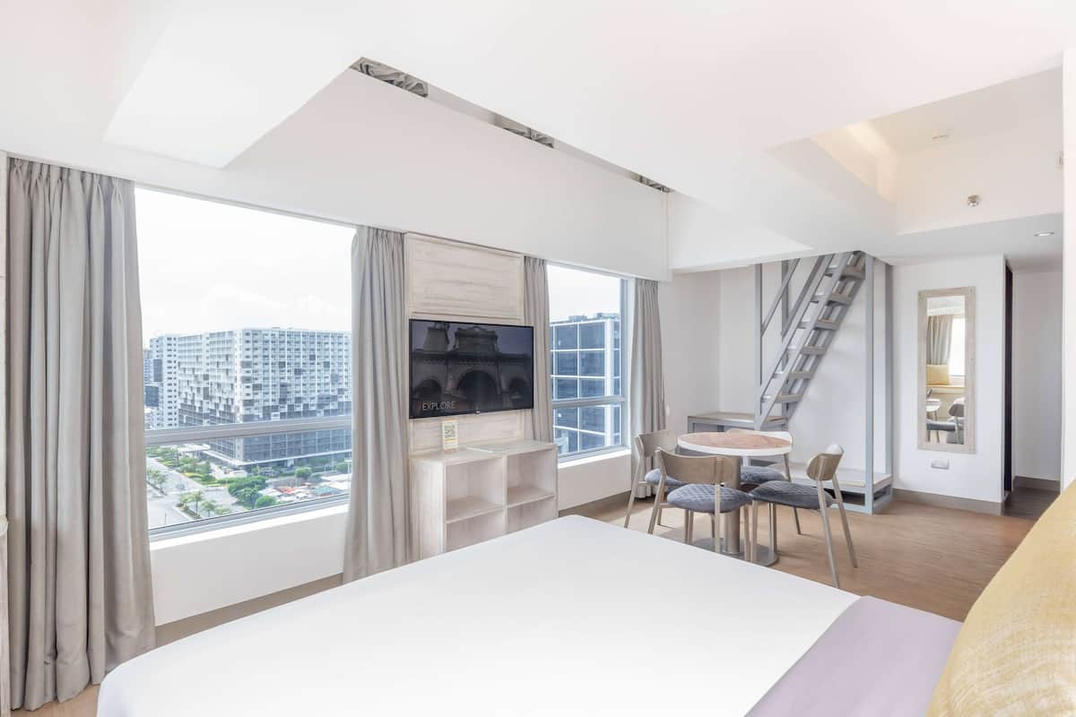 Een moderne hotelkamer met een groot bed, een aan de muur bevestigde televisie, een zithoek met tafel en stoelen, en uitzicht op de stad door grote ramen.