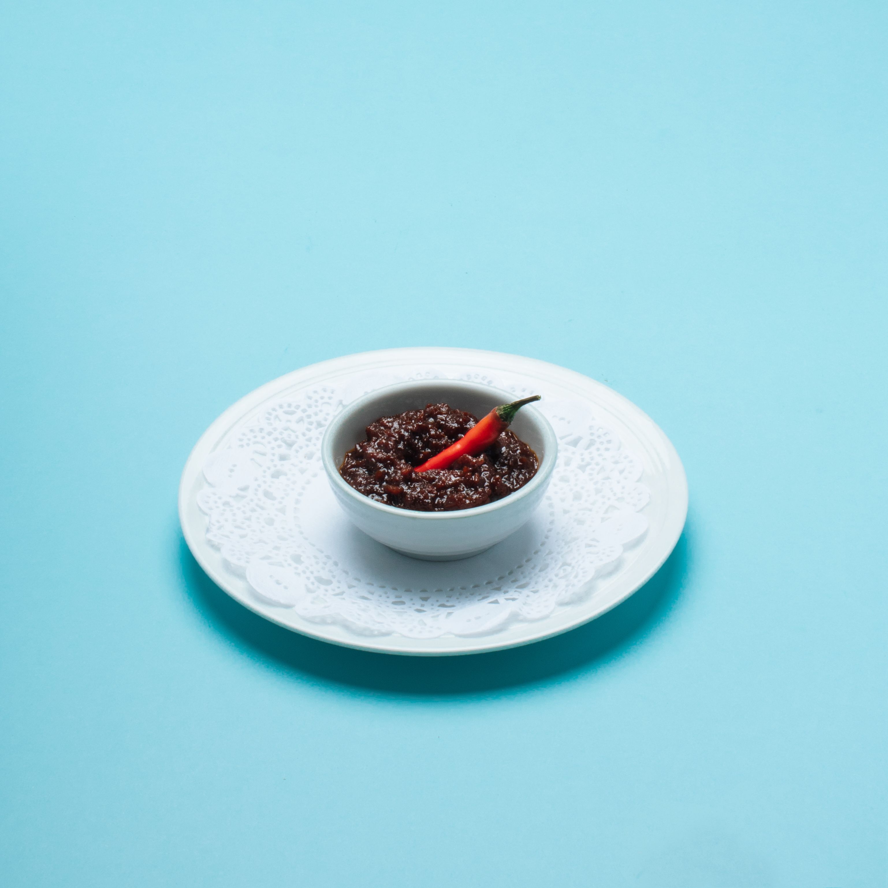 Un bol contenant des aliments de couleur foncée, avec un piment rouge, posé sur une assiette blanche, elle-même placée sur une surface bleue.