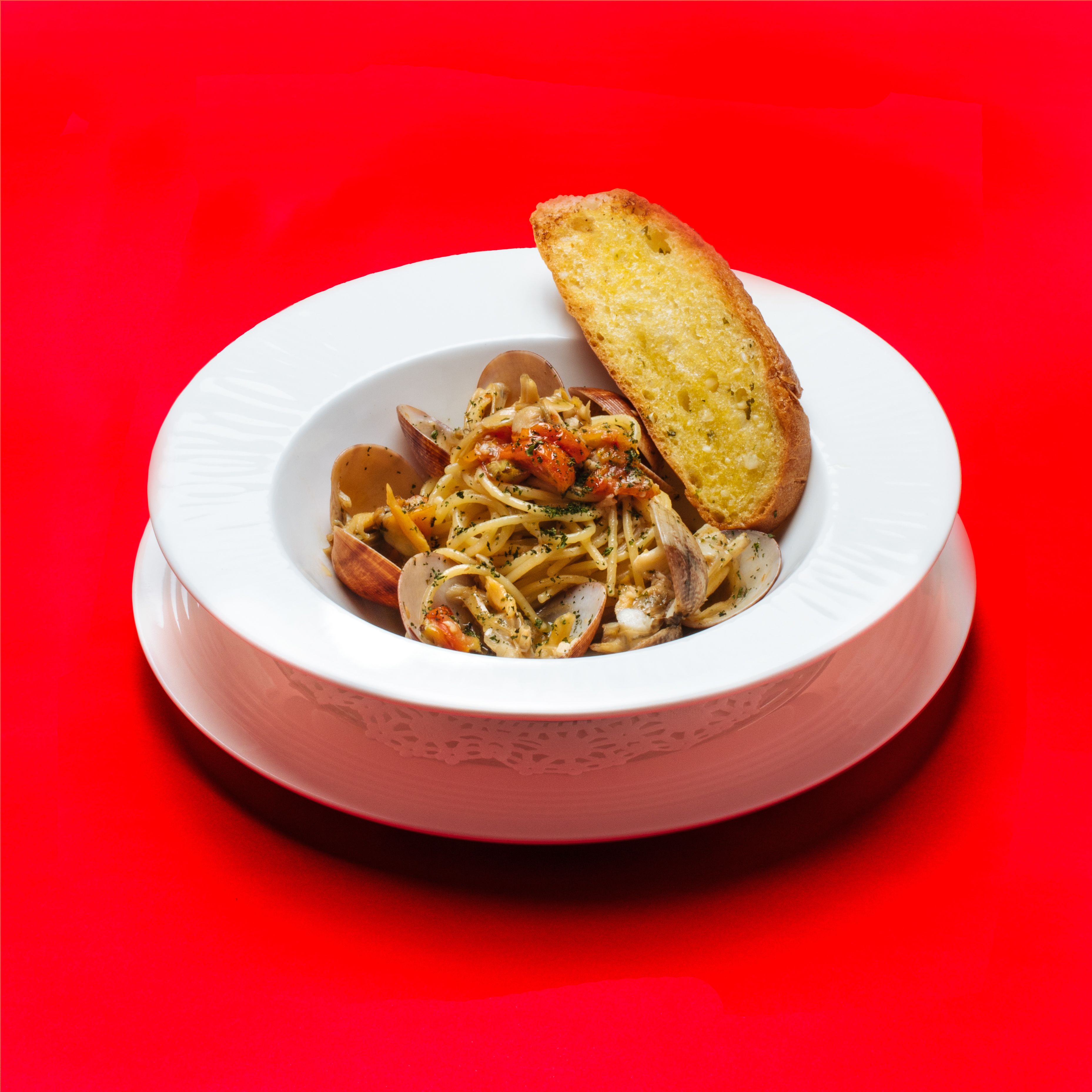 A white bowl with pasta and clams, accompanied by a piece of toasted bread on a red background.