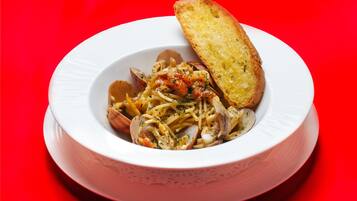 A white bowl with pasta and clams, accompanied by a piece of toasted bread on a red background.