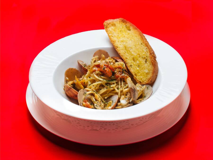A white bowl with pasta and clams, accompanied by a piece of toasted bread on a red background.