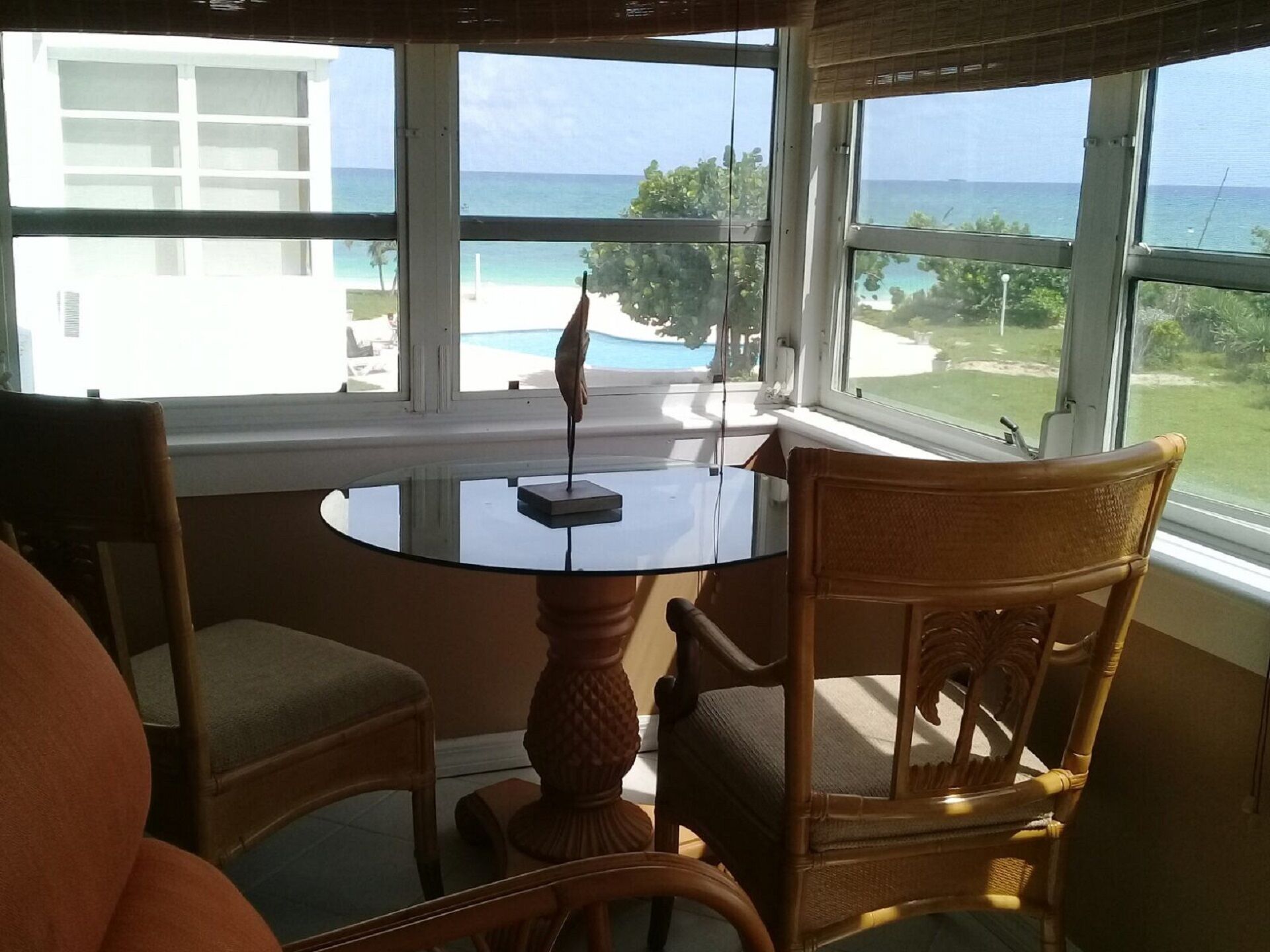Una habitación con una mesa de vidrio y dos sillas de mimbre, una ventana con vista al océano y un jardín.