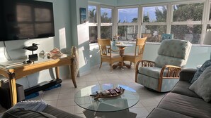 Une pièce lumineuse, avec un téléviseur, une table basse en verre, un canapé et un coin repas comprenant des chaises et une table.