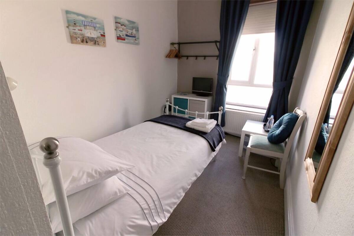 A small, neatly arranged bedroom with a single bed, a bedside table with a lamp, a chair, and a TV.
