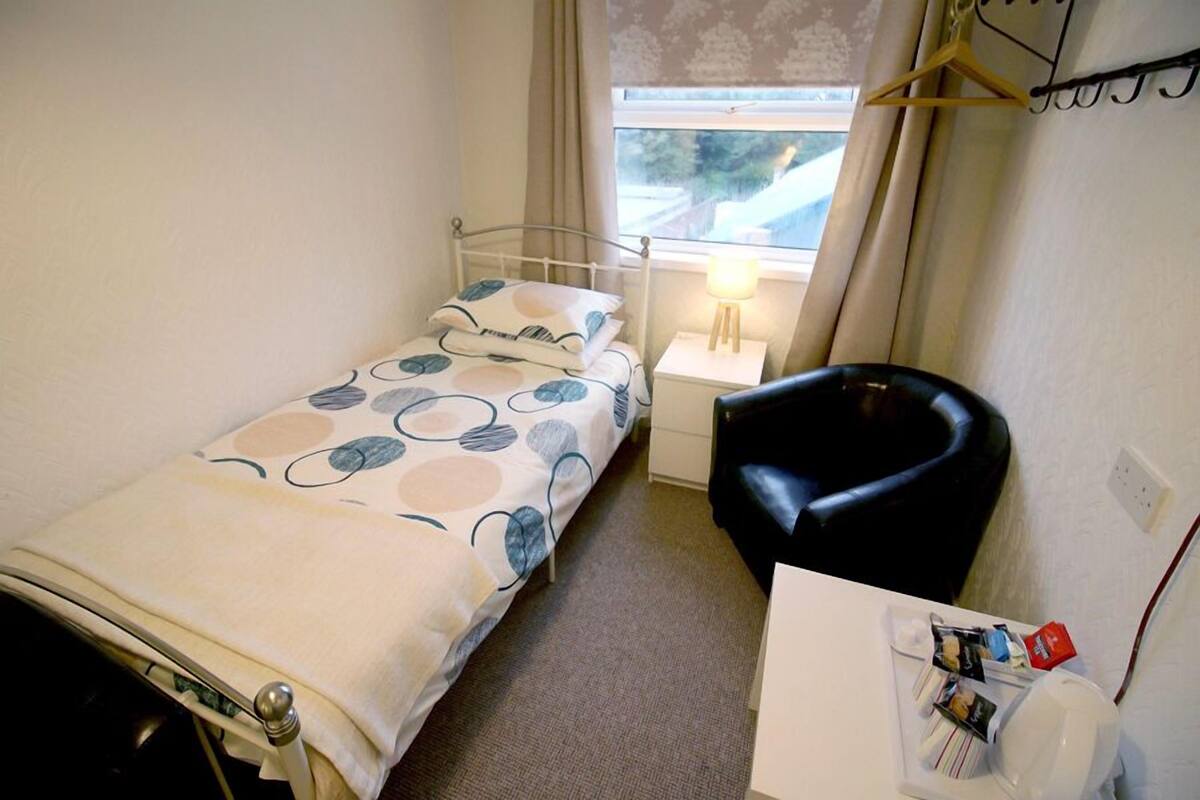 A small, compact room with a single bed, a bedside table with a lamp, a black chair, and a window with curtains.