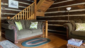 L’intérieur d’une cabane en rondins, avec un escalier en bois, deux canapés ornés de coussins à motifs, un tapis et une boîte posée sur une table.