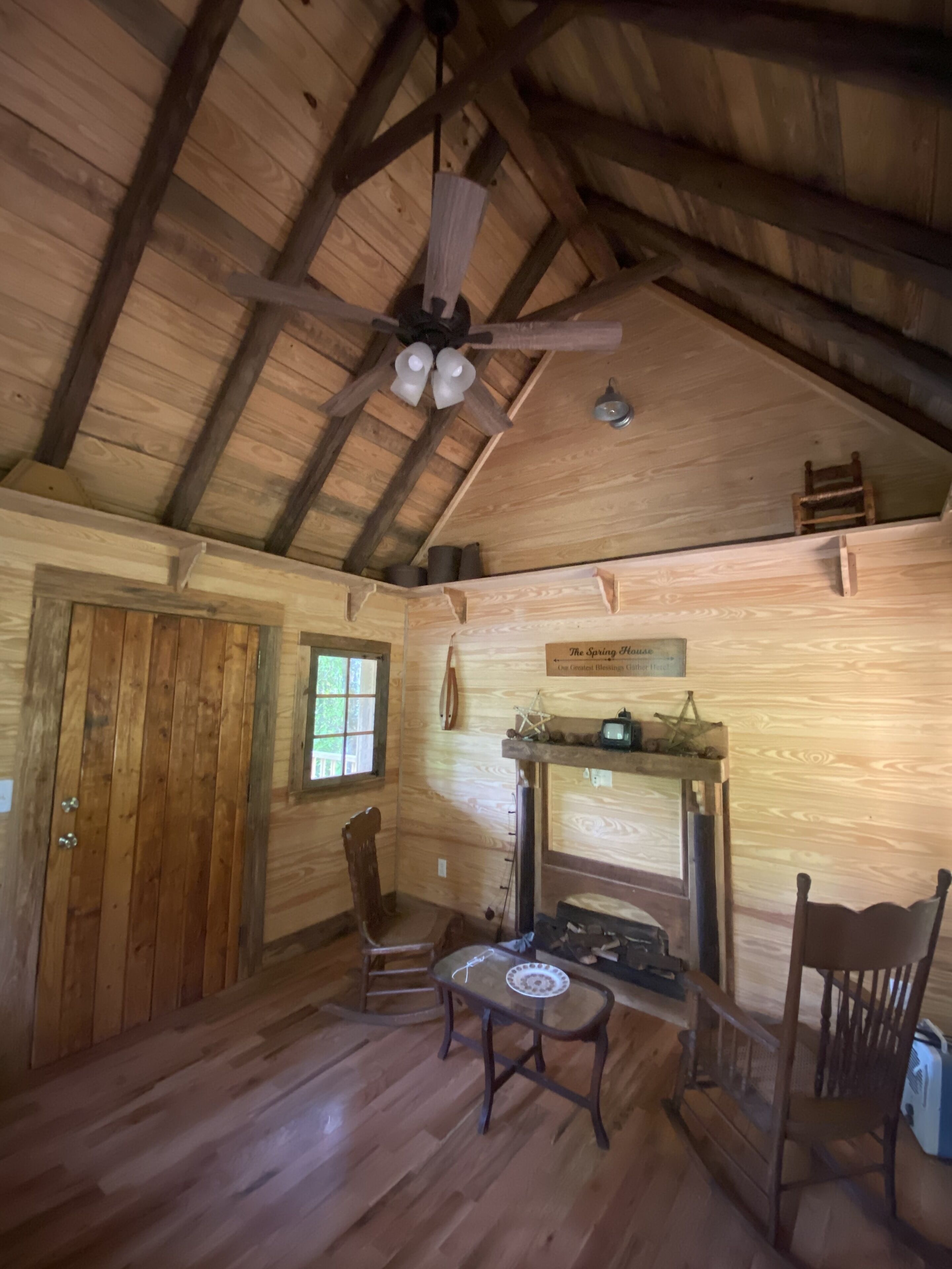 A rustic wooden interior with a sloped ceiling, a ceiling fan, a fireplace, and wooden furniture.