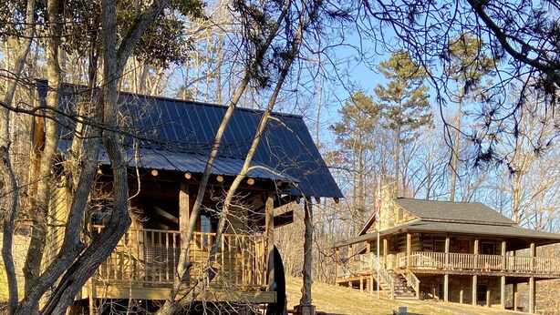 Un chalet rustique doté d’une terrasse en bois et de fondations en pierre, entouré d’arbres et bordé d’un petit étang.