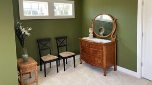 Quarto com cômoda de madeira, duas cadeiras, um vaso com flores e janela com vista para o verde.