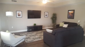 A living room with a dark-colored sofa, a white chair, a flat-screen TV mounted on the wall, a floor lamp, and a potted plant. - Estancia Resort! New beauty in St George with heated pool and pickleball (Saint George)