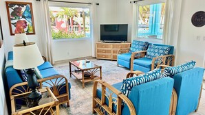 A living room with blue furniture, a television, and a large window with curtains. - Sunset Beach "C1" -From the Veranda to the beach! Free Bikes, Kayaks, & More 🏖 (San Pedro Town)
