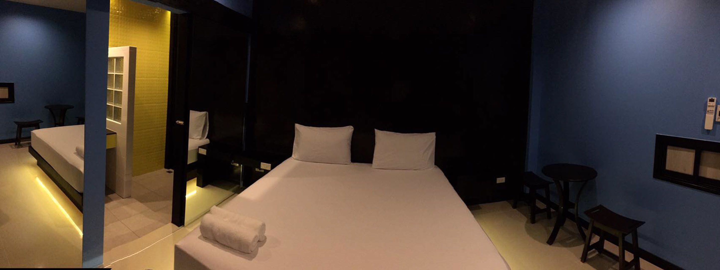Habitación de hotel con una cama grande, una mesita de noche, un taburete pequeño, un espejo y un baño con inodoro y lavabo.