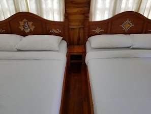 Uma cama de casal com cabeceira de madeira e detalhes florais, uma mesinha de cabeceira de madeira e uma janela com cortinas brancas.