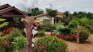 Un comedero para pájaros con un ave posada, rodeado de vegetación exuberante y una casa al fondo.