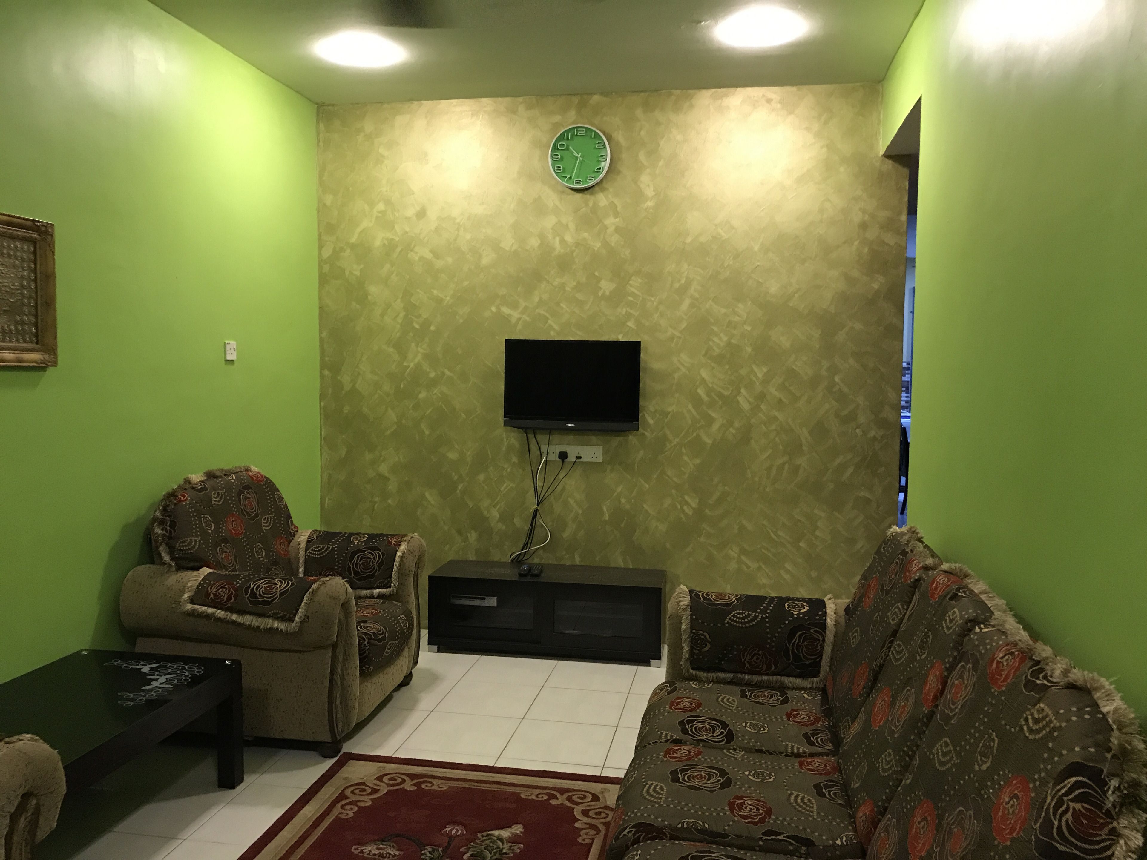 Uma sala de estar com paredes verdes, um relógio, uma TV de tela plana, dois sofás estampados e uma mesa de centro.