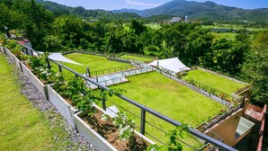 A view of a well-maintained lawn with a tennis court, surrounded by trees and hills in the background. - Hotel on Hilltop (Kathu)