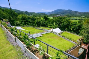Vista de un césped cuidado con una cancha de tenis, rodeado de árboles y colinas al fondo.