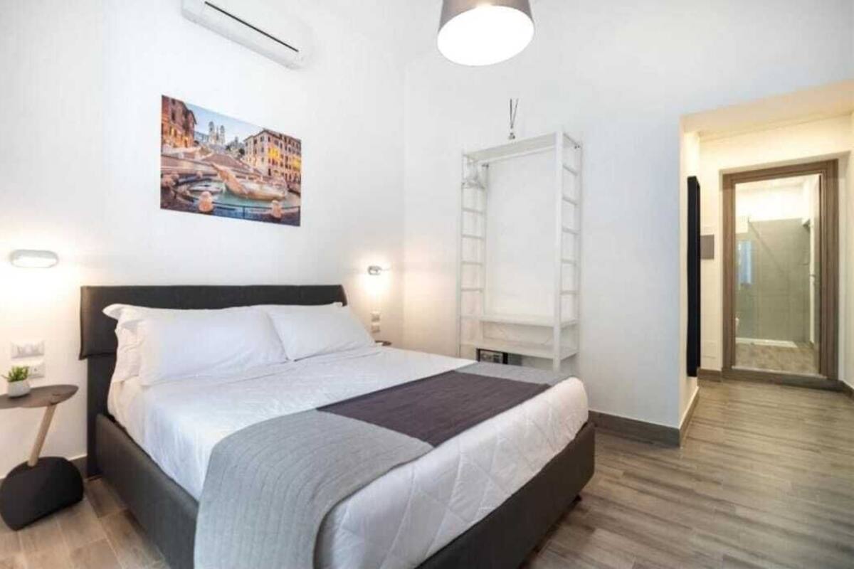 A bedroom with a bed, bedside tables, a wall-mounted air conditioner, a painting, and a door leading to another room.