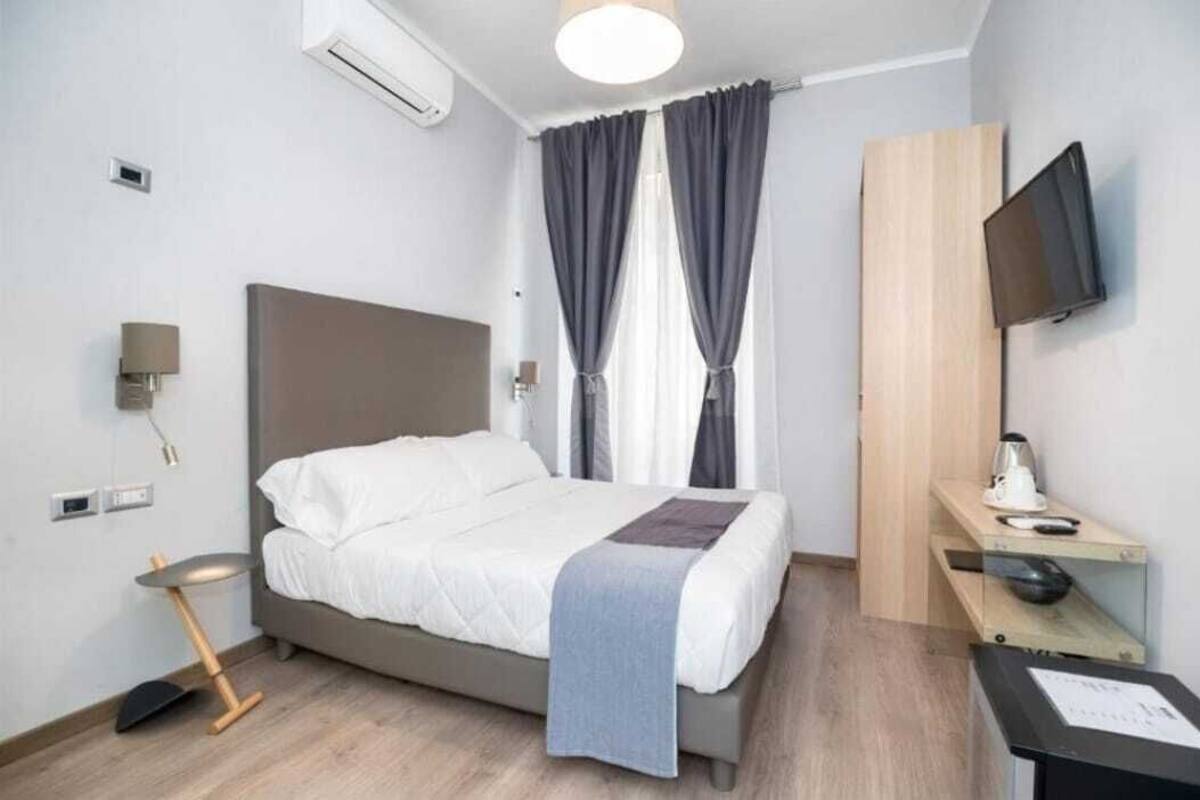 A modern hotel room with a bed, a desk with a flat-screen TV, a lamp, and a window with curtains.