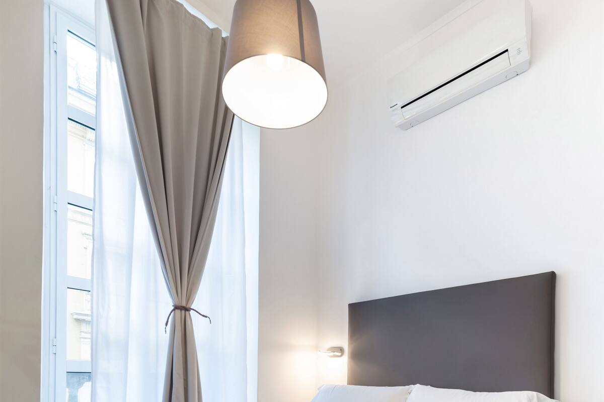 A modern bedroom with a large bed, a hanging light fixture, an air conditioning unit, and a view through a window with curtains.