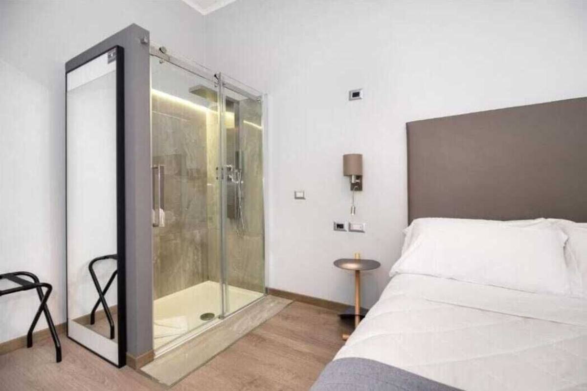 A modern hotel room with a glass-enclosed shower, a bed with white linens, a round side table, and a wall-mounted lamp.