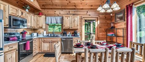 A wooden cabin with a kitchen featuring a microwave, stove, and dishwasher. There is a dining table with chairs and a chandelier.