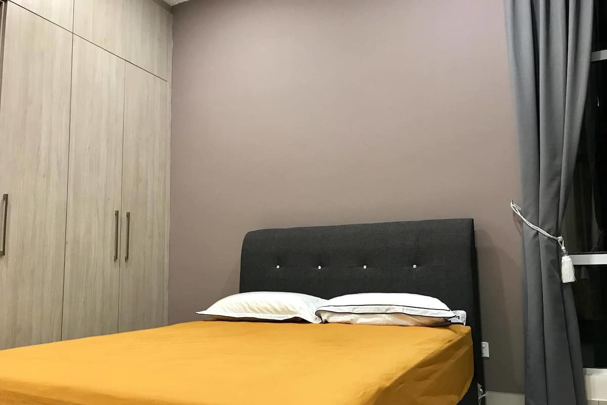 A bed with a dark headboard, a mustard-colored bedspread, and white pillows. There is a wardrobe with light wood finish and metal handles. The room has a plain wall and a dark curtain on the right side.