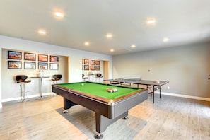 Une table de billard recouverte d’un drap vert, deux queues de billard et un jeu de billes. Il y a trois tableaux encadrés au mur et deux tabourets de bar.