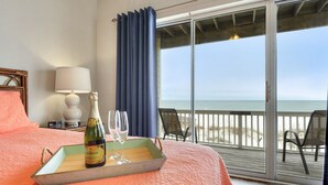 A hotel room with a bed, a tray with a bottle and glasses, a lamp, and a view of the beach through a sliding glass door.
