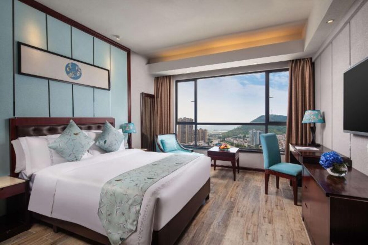 Ein Hotelzimmer mit einem großen Bett, einem Schreibtisch, einem Stuhl und einem Fernseher. Das Zimmer verfügt über ein großes Fenster mit Blick auf die Stadt.