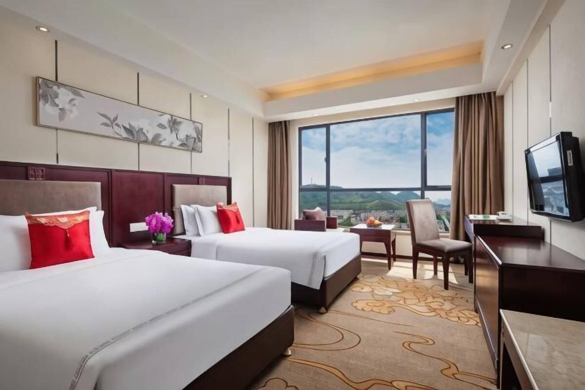 Habitación de hotel con dos camas, un escritorio, un televisor y un ventanal con vistas.