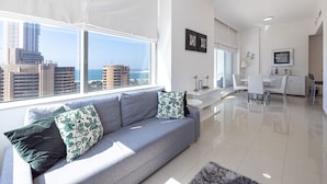 A modern living room with a grey sofa, a dining area with white chairs, and a view of the cityscape through large windows. - DHH - Botanica Tower (Dubai)
