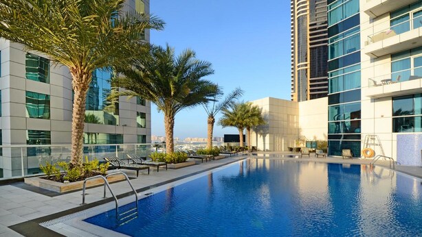 A rooftop swimming pool with lounge chairs and palm trees, surrounded by modern buildings. - DHH - Botanica Tower (Dubai)