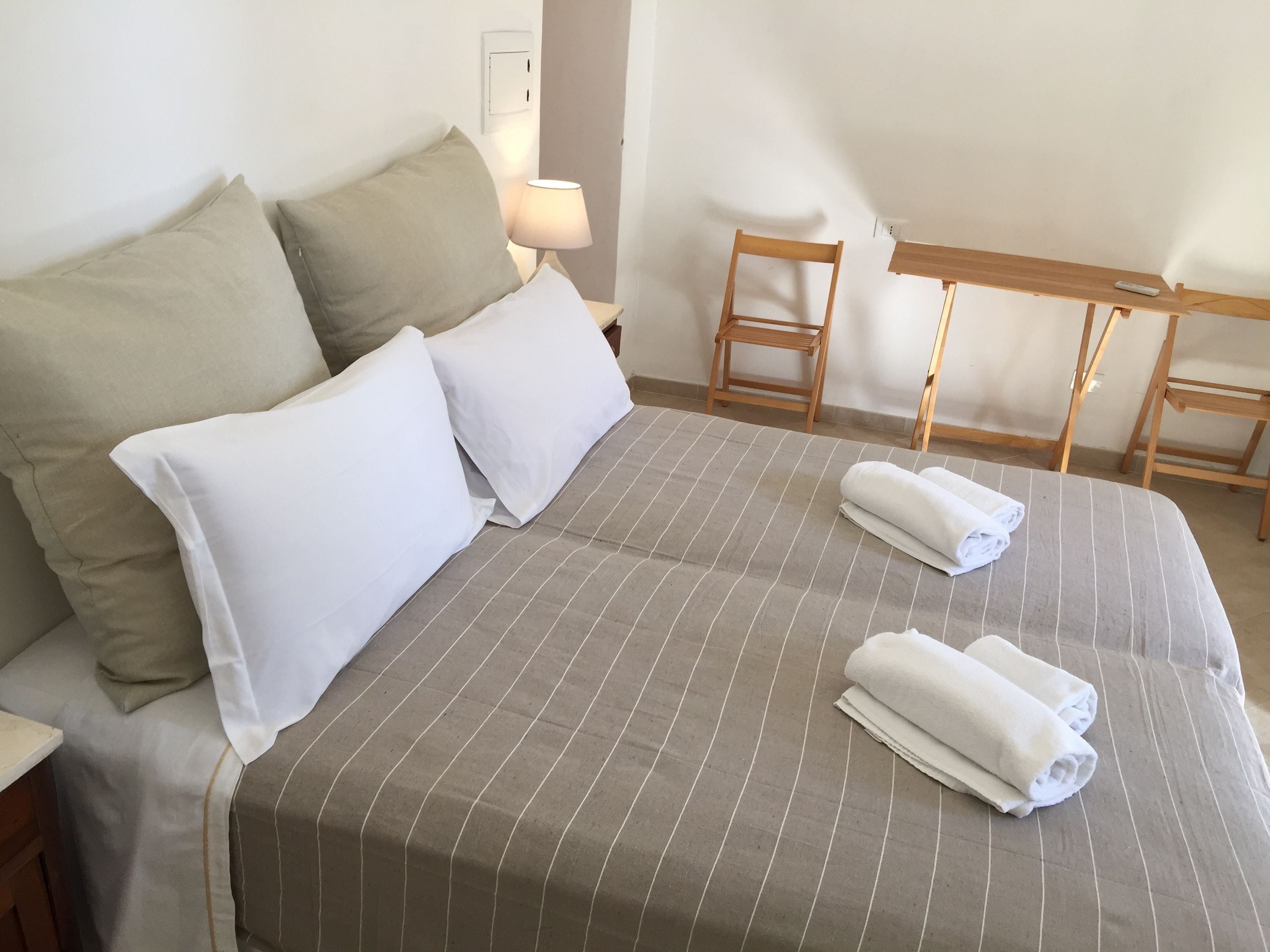 A neatly made bed with striped bedding and two rolled towels, a wooden chair, and a small table with a lamp in a simple, clean room.