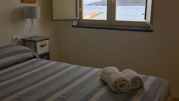 Una cama con colcha a rayas, una toalla enrollada y una ventana con vista al mar.