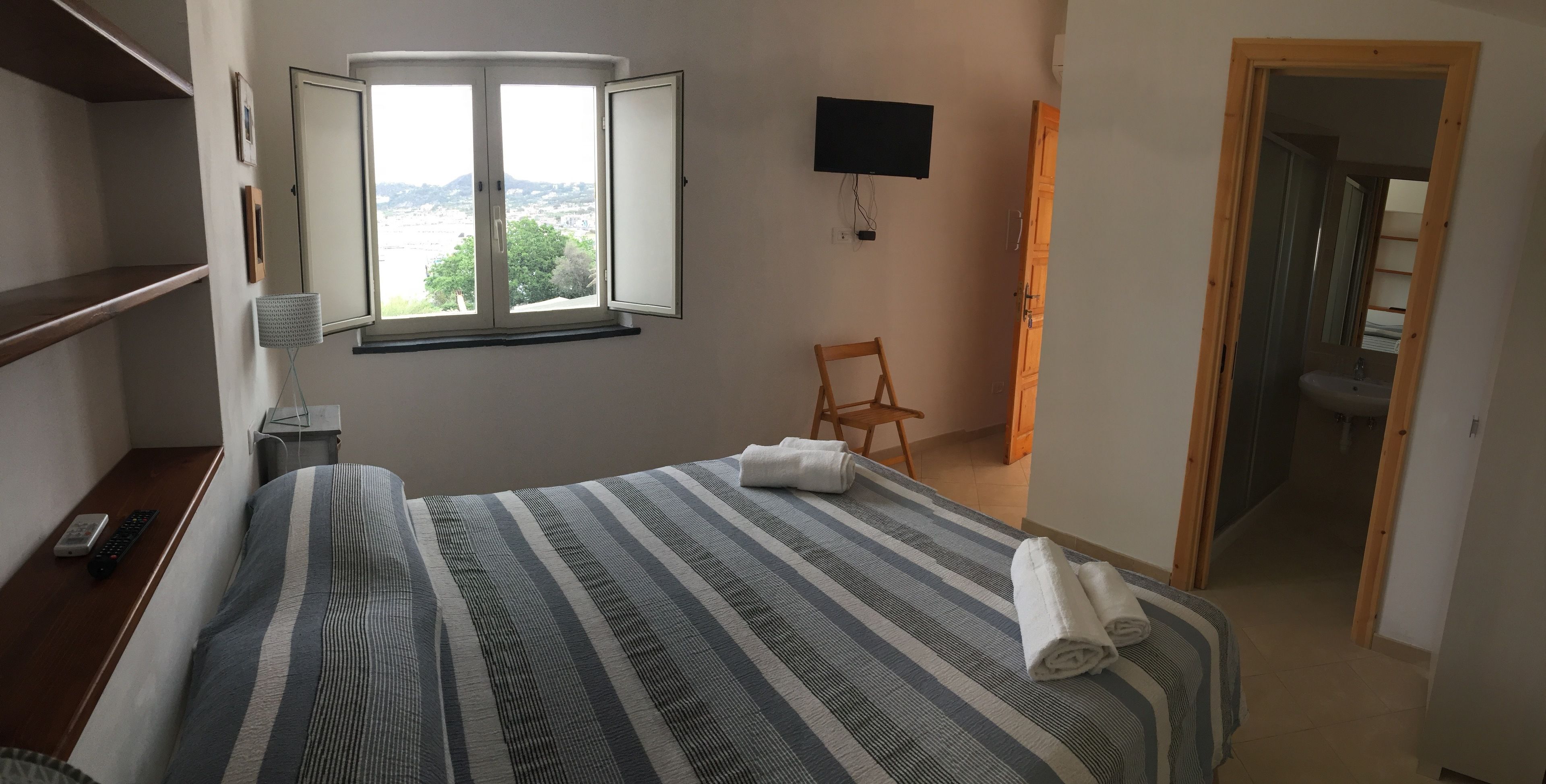 A hotel room with a bed, a TV mounted on the wall, a window with a view, a chair, and a bathroom visible through an open door.