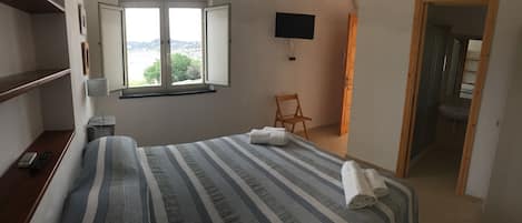 Une chambre d’hôtel avec un lit, un téléviseur fixé au mur, une fenêtre donnant sur un paysage agréable, une chaise et une salle de bain visible à travers une porte ouverte.