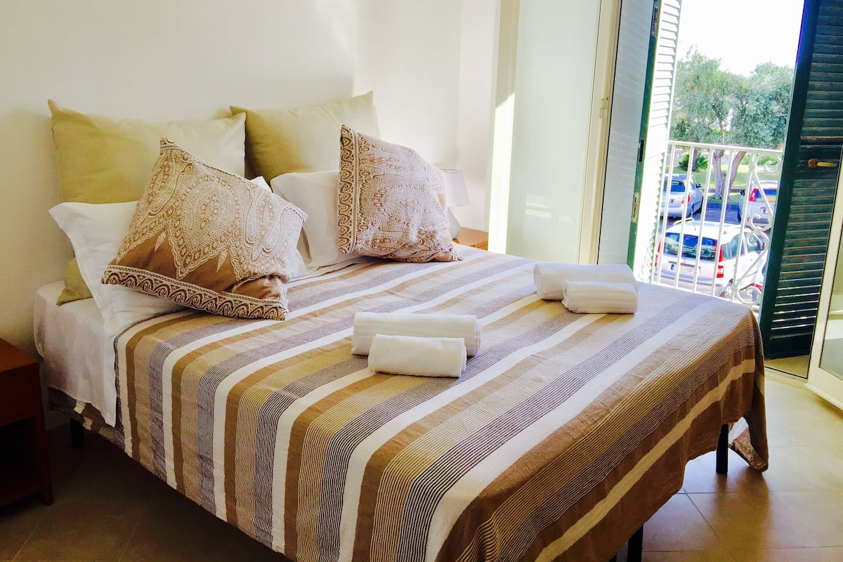 A neatly made bed with striped bedding and decorative pillows, a bedside table with a lamp, and a view of parked cars through a glass door.