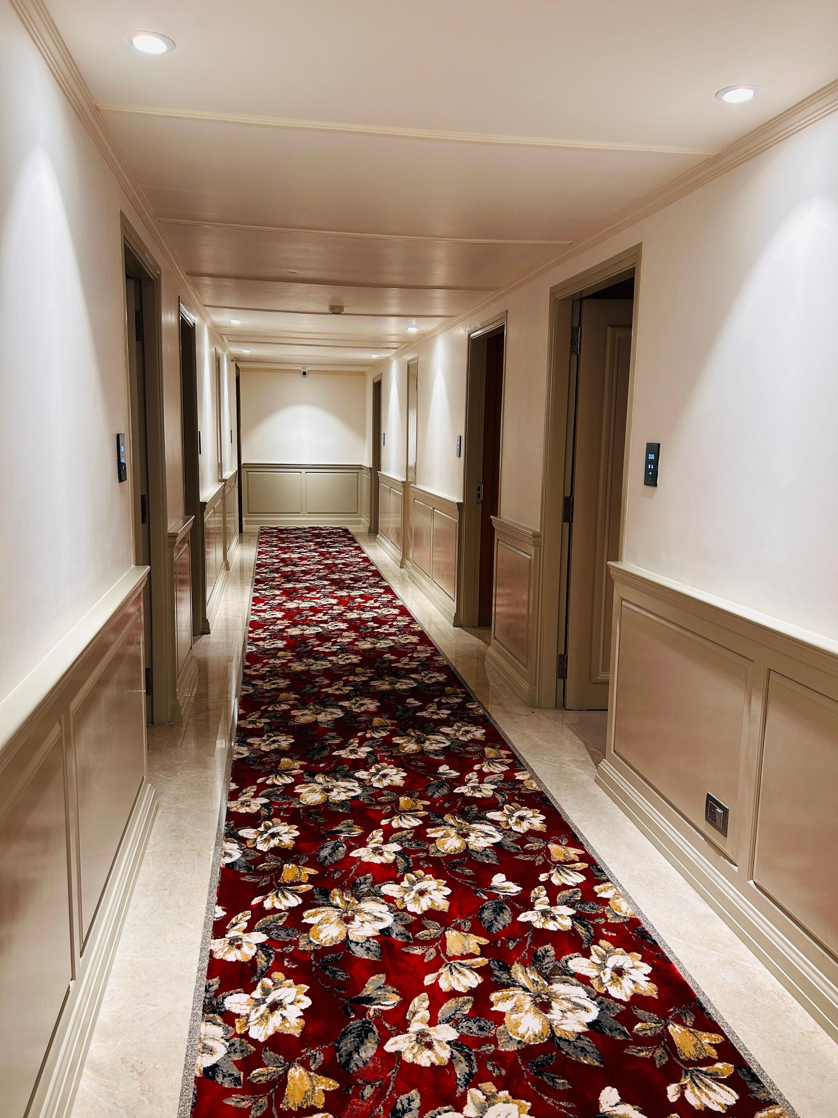 A hotel hallway with a red floral carpet, cream-colored walls, and a ceiling with recessed lighting.