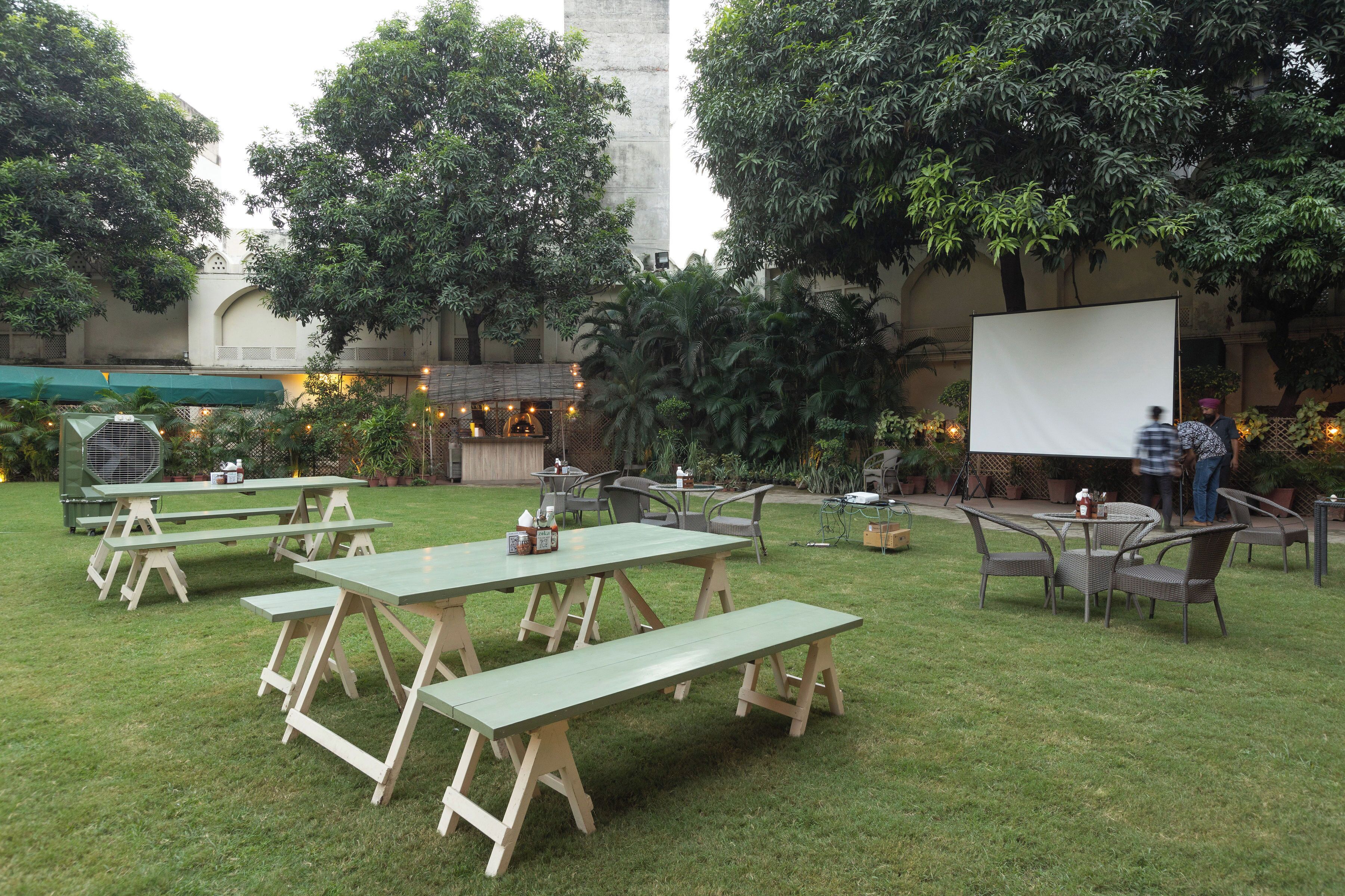 An outdoor patio area with picnic tables and chairs, surrounded by lush greenery and a large white screen.